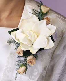 This corsage contains flowers such as gardenia, white spray 	roses and greens.